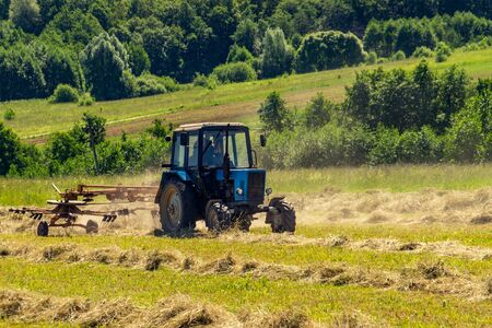 An old tractor turns over the mowed hay on a Sunny summer morning for better drying. Fodder for cows for the winter.の写真素材