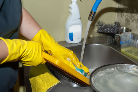 washing dishes under running water close-up. Womens hands in yellow household gloves clean dishes before washing in the dishwasherの写真素材