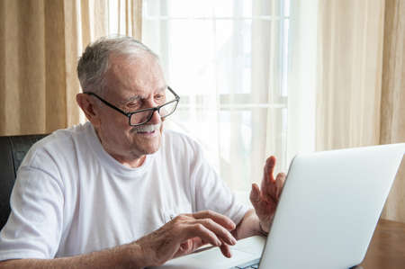 Older man at home in front of a laptop. A gray-haired old man with glasses works at home in front of a laptop. The concept of communication of old people.の写真素材