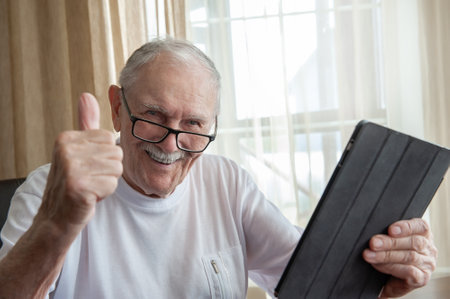 Happy elderly man at home with laptop. A gray-haired old man with glasses is happy and gestures that everything is OK. The concept of communication between older people.の写真素材