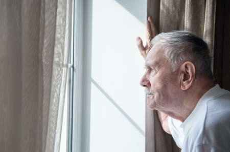 gray-haired old man, forced to remain in quarantine due to the virus, looks out the window with a disconsolate expression. Loneliness of old people. Stay home in the pandemic.の写真素材
