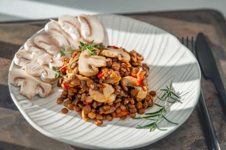 lentils with fried sliced mushrooms and vegetables. Vegan food - stewed carrots, peppers, onions and lentils with fried mushrooms. A dish of lentils on a beautiful light plate with a black fork and knife. Close-up.の写真素材