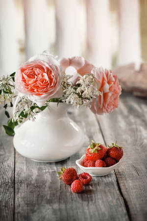 Ripe strawberries in a white cup on the table in the morning. Summer breakfast. Still life of red strawberries and a bouquet of roses with, copy spaceの写真素材