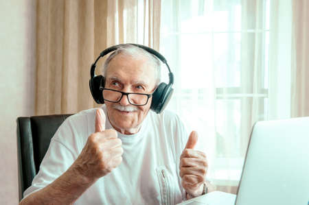 portrait of an elderly man with headphones. Positive human emotions, facial expression. A smiling elderly man using a laptop at home, listening to his favorite music and looking for information on the Internet, a happy man makes a video call.の写真素材