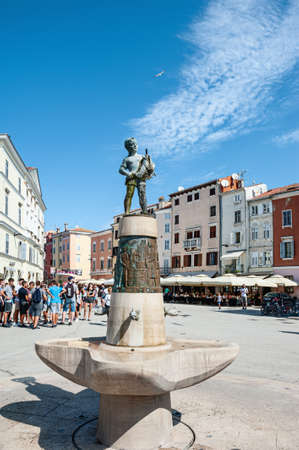 ROVINJ, CROATIA-AUGUST 30, 2018: Tourists walk by the Fountain with a sculpture of a boy and a fish and a clock Tower on the Picturesque Town Square in Rovinj, Croatiaのeditorial素材