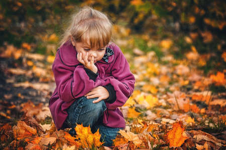 little girl in the fall. 5-year-old child sits thoughtfully among the autumn leaves. real childish emotionの写真素材
