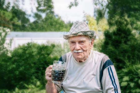 large portrait of a gray-haired old man in a hat with a glass of dark drink. man with a glass mug is sitting in the garden.の写真素材