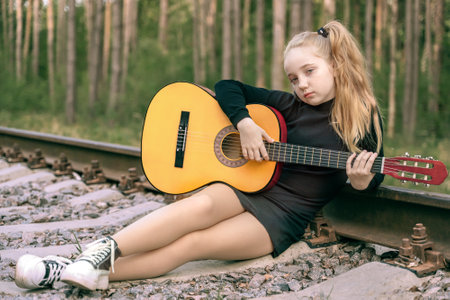 A 13-year-old teenager with a guitar. The girl travels with a guitar. The girl sat down to rest on the rails in the forest. Inspiration, talent.の写真素材