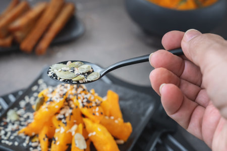 Sesame and pumpkin seeds in a spoon for sprinkling the pumpkin dish. Pumpkin pieces on a black flat...