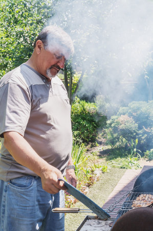 An elderly man in the countryside cooking a barbecue. A gray-haired man enjoys the nice weather and cooks food on an open fire. Smoke over the barbecue.の写真素材