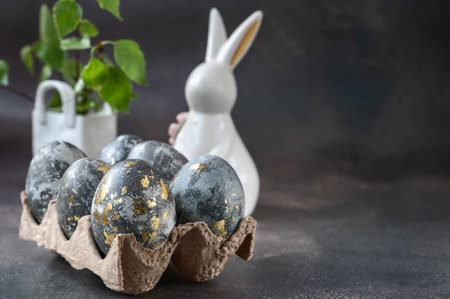 Spring Easter still life. A white rabbit and eggs painted with a gold pattern in an eco-friendly stand made of recycled cardboard on a gray background. Easter decor on the table. Copy spaceの写真素材