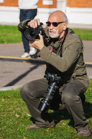 October 1, 2016, Moscow. Cossack gathering. gray-haired elderly photographer smiles cheerfully with a camera in his hand. cheerful old photographer with a beard and glasses takes pictures of people in the parkのeditorial素材