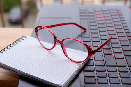 Stylish glasses in a red frame close-up. Nearby on the laptop keyboard lies an open notepad with space for text. .の写真素材