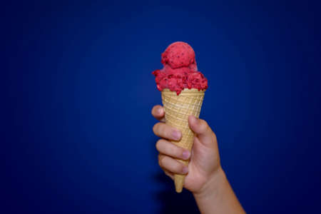 Berry red ice cream close-up. A child's hand holds a delicious ice cream cone on a blue background.の写真素材