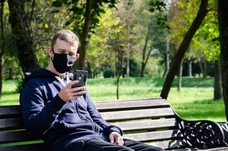 Caucasian man in protective medical mask making video call on phone and sitting on bench in the park during coronavirus outbreak with copy space. New reality. Long distance relationship concept.の写真素材