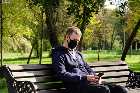 Caucasian man in protective medical mask working on phone and sitting on bench in the park during coronavirus outbreak. New reality. Freelancer concept.の写真素材