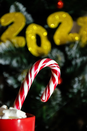Close up red cup with sweet caramel cane and marshmallows on the background of Christmas tree with the numbers 2021. New year concept. 2021 happy new year eve background with golden colored numbers.の写真素材