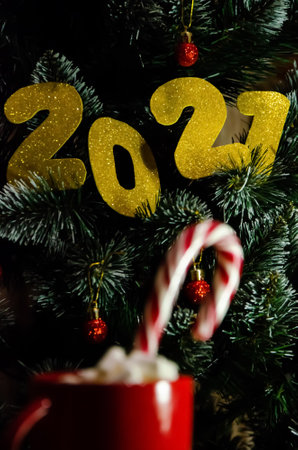 Blurred red cup with sweet caramel cane and marshmallows on the background of Christmas tree with the selective focus on numbers 2021. New year concept. 2021 happy new year eve background with golden colored numbers.の写真素材