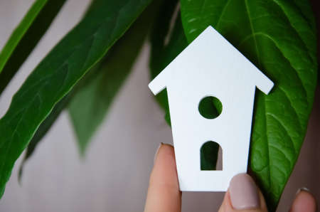 Own housing concept. Female hand holds white decorative house on background of green leaves with copy space. Stay home.の写真素材
