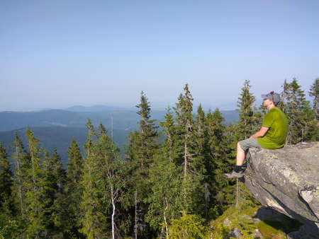 Selective focus caucasian man is resting on stone against background of forest, mountains and sky....