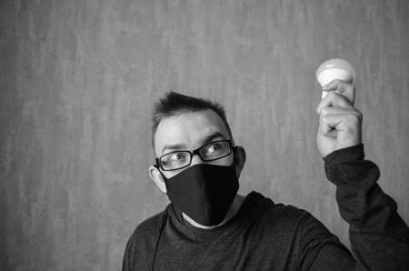 Caucasian man in protective mask with bulb in his hand on gray background with copy space. Monochrome photo with man. Concept of creating new ideas. Portrait of excited male.の写真素材