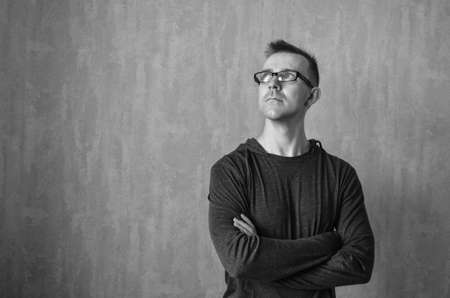 Caucasian man in glasses looking up and standing with folded arms on gray background. Monochrome portrait of pondered or dreamed male with copy space. Thinking concept.の写真素材