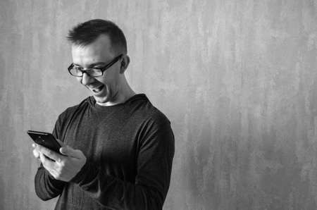 Caucasian man in glasses looks at phone and rejoices on gray background. Monochrome portrait of delighted male with copy space. Concept of joy and success.の写真素材