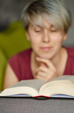Selective focus on book read by smiling caucasian woman with short haircut on gray sofa. Concept of...