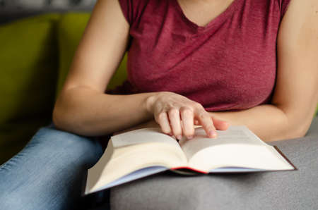 Selective focus on book read by caucasian woman in burgundy T-shirt and jeans sitting on gray sofa...