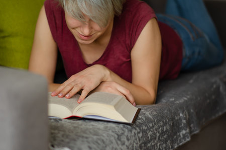 Selective focus on book read by caucasian woman with short haircut in burgundy T-shirt and jeans...