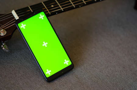 Selective focus on phone with greenscreen of chromakey mockup with tracking markers near the guitar with copy space. Green screen and chroma key. Online guitar courses.の写真素材