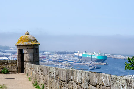 Ancient stone watchtower with view of busy maritime port, large cargo ship and marina under blue summer sky. Perfect for themes of travel, history, and industry.の写真素材
