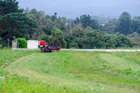 A person operates a riding lawn mower, maintaining a green field surrounded by trees and mountains. Rural work scene, outdoor activity, and landscape maintenance are depicted.の写真素材
