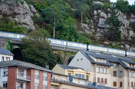 A modern passenger train travels on a viaduct through lush greenery and rocky hills, passing over a...