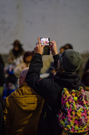 A crowd watches a vibrant outdoor street performance as a person in a beanie and colorful backpack captures the moment on a smartphone, showcasing urban lifestyle.の写真素材