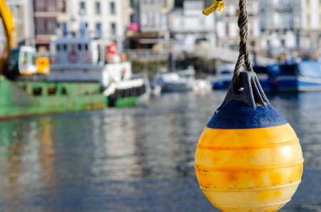 Bright yellow buoy hanging over calm marina water, capturing the atmosphere of a busy harbor with moored boats and a lively coastal town in the background.の写真素材