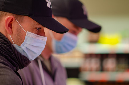 Two adult men in medical masks and black caps shop together in a brightly lit supermarket amid...