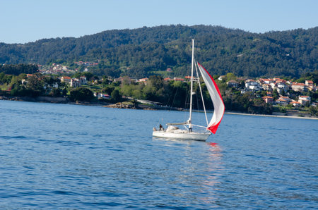 Scenic view of a sailboat cruising on calm blue water near a picturesque village by the forested...