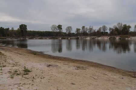 Blue river slow flowing across the green meadow and yellow sand with reflections of trees in the...