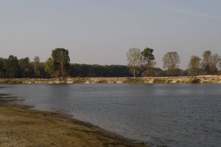 Blue river slow flowing across the green meadow and yellow sand with reflections of trees in the...