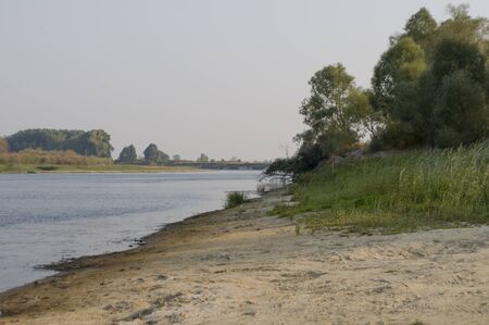 Blue river slow flowing across the green meadow and yellow sand with reflections of trees in the...
