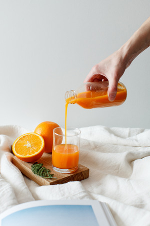 The girl pours an orange fresh from a bottle into a glass. There is an orange on the wooden board and magazine. Healthy breakfast in bed, daylight, vertical shot.Healthy breakfast in bed, daylight, vertical shot.の写真素材