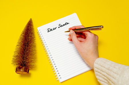 Female hand writing letter to Santa on yellow background. Golden Christmas tree and dear Santa handwriting. Christmas concept.の写真素材