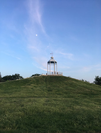 A white gazebo standing high on the hillside covered with fresh green grass under the blue spring sky with white wing-like clouds and moon; a vertical photoの写真素材
