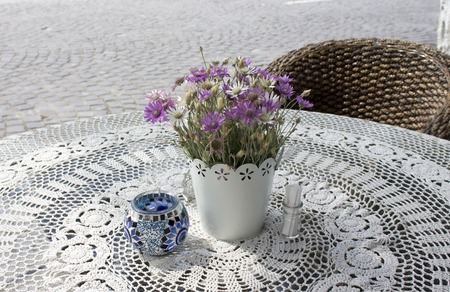 Photo of cozy cafe table on paving stone street in the sunny summer morning with beautiful flowers on lace tablecloth and wooden armchair.の写真素材