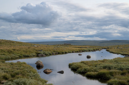 A peaceful scene from the Norwegian countryside featuring a calm river winding through grassy...