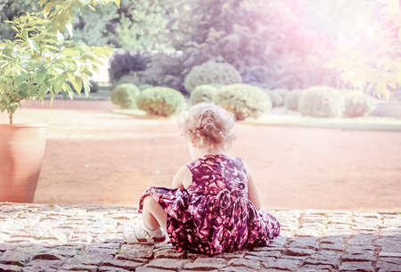 Blurred view of a little curly girl who sits on a stone walkway in a summer park. High quality photoの写真素材