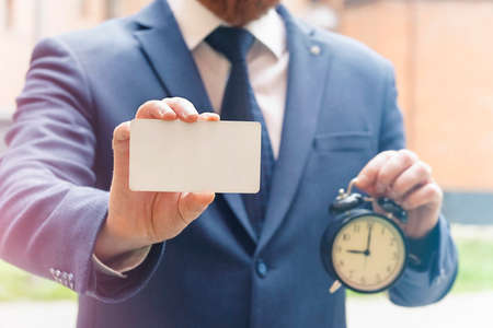a man in a suit holds a blank card in one hand and an alarm clock that shows 9 am in the other hand....