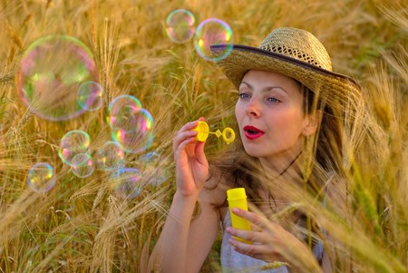 Girl in wheat field in white dress and hat  blowing soap bubbles. Selective focus.の写真素材