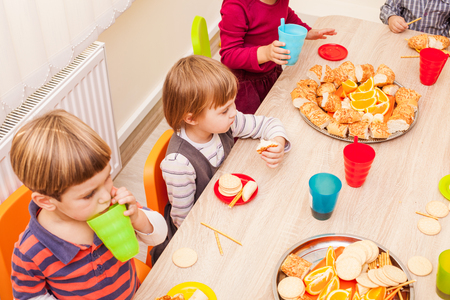 Children are sitting at the table with lunch and eating fruits and cakesの写真素材